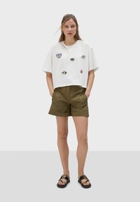 Camiseta blanca corta con parches coloridos de un ojo y un corazón, combinada con pantalones cortos de color verde oliva y sandalias negras con acentos dorados.