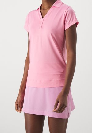 Femme portant un haut rose à manches courtes avec col et une jupe rose clair sur un fond clair uni, face à l'avant.