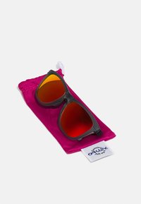 Occhiali da sole neri con lenti rosse, poggiati su un sacchetto di satin rosa che presenta un'etichetta bianca con un logo blu in basso.