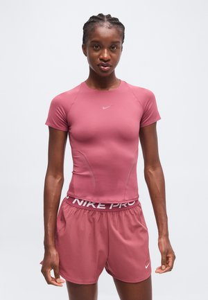 Kobieta ubrana w dopasowaną różową sportową koszulkę i pasujące do niej szorty z logo Nike, stojąca na jednolitym jasnym tle.