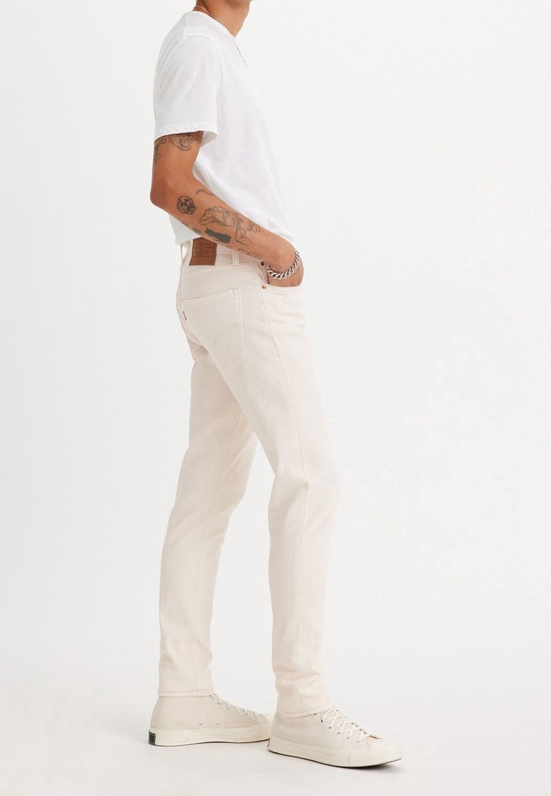 Personne portant un jean slim blanc, un t-shirt blanc et des baskets blanches, debout de profil avec une main dans la poche, sur fond uni.