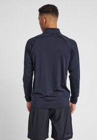 Maglietta atletica a maniche lunghe blu navy con colletto alto e maniche raglan. Il retro presenta una texture liscia e sottili accenti con il logo.