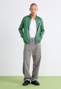 Homem a usar uma jaqueta verde com fecho éclair, uma t-shirt branca, calças de cotim cinzentas e sapatos pretos, de pé com as mãos nos bolsos sobre um chão verde.