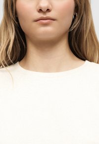 Νεαρή γυναίκα με ανοιχτά καστανά μαλλιά που φοράει λευκό μπλουζάκι με στρογγυλή λαιμόκοψη, απεικονιζόμενη από τον λαιμό έως τους ώμους μπροστά σε απλό φόντο.