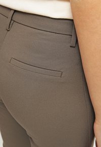 Närbild av en persons höft iförd åtsittande taupebyxor med en bakficka och en beige topp instoppad.