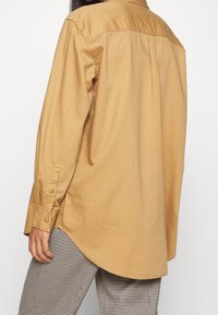 Camicia a maniche lunghe con bottoni in tessuto marrone chiaro, dotata di una spalla posteriore, orlo curvo e spacchi laterali, abbinata a pantaloni a scacchi.