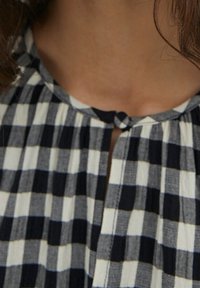 Mustavalkoinen ruutukangas, jossa on pleatattu muotoilu; siinä on pyöreä kaula-aukko ja pieni, solmittu nappikiinnitys edessä.