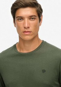 Ung mand med kort brunt hår og grønne øjne iført en olivengrøn t-shirt med rund hals mod en ensfarvet lys baggrund.