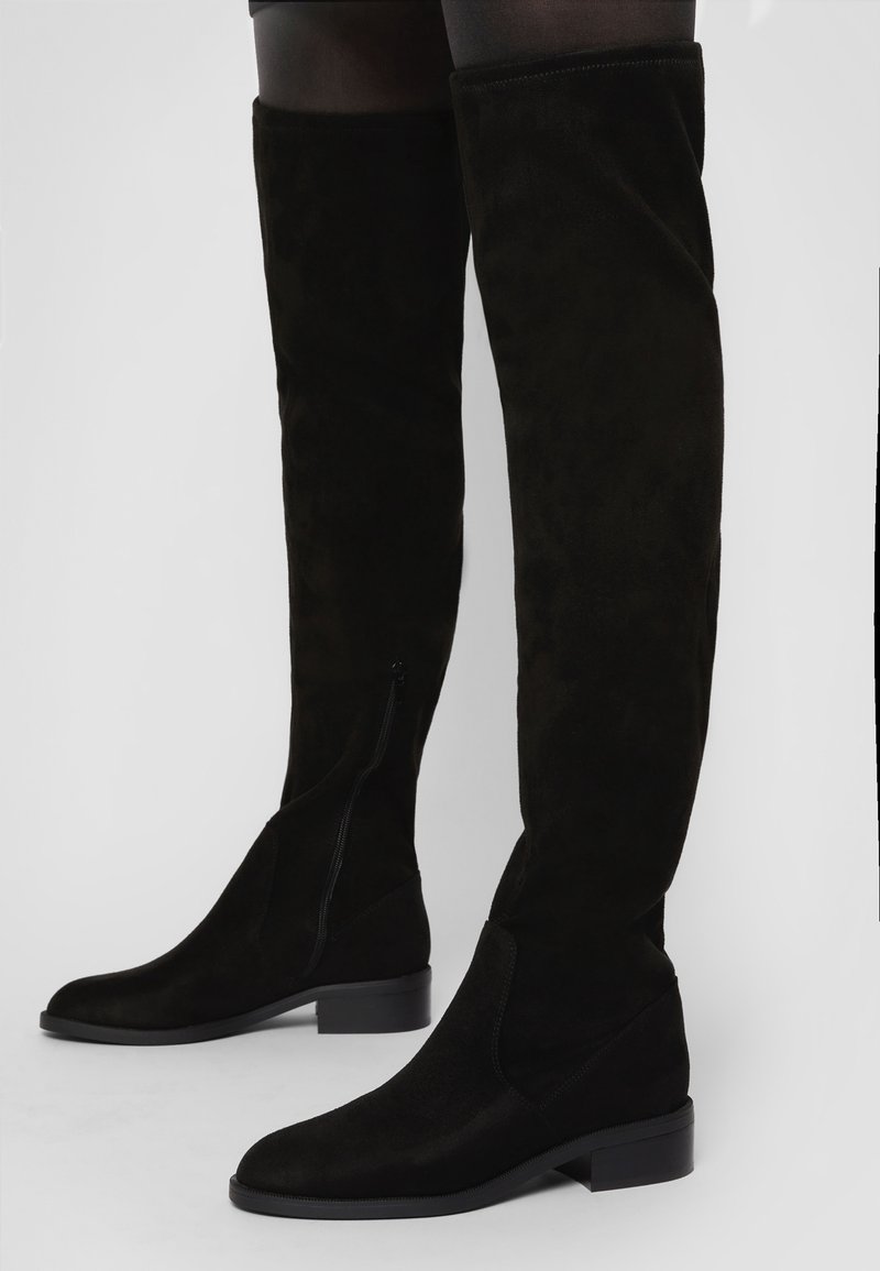 Bottes hautes en daim noir jusqu'aux cuisses avec talons carrés bas portées avec des collants noirs transparents sur un fond uni.