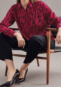 Blusa a fantasia rossa e nera con chiusura a bottoni, abbinata a pantaloni neri a tre quarti e scarpe con tacco nere, seduta su una sedia di legno.