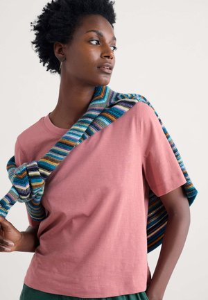 T-shirt rosa a maniche corte realizzato in tessuto morbido, indossato con un maglione lavorato a maglia a righe colorate che pende sulla spalla.
