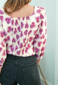 Blusa a maniche lunghe con base bianca, caratterizzata da un motivo floreale paisley rosa e rosso. Design arricciato sul retro, abbinata a jeans neri a vita alta.