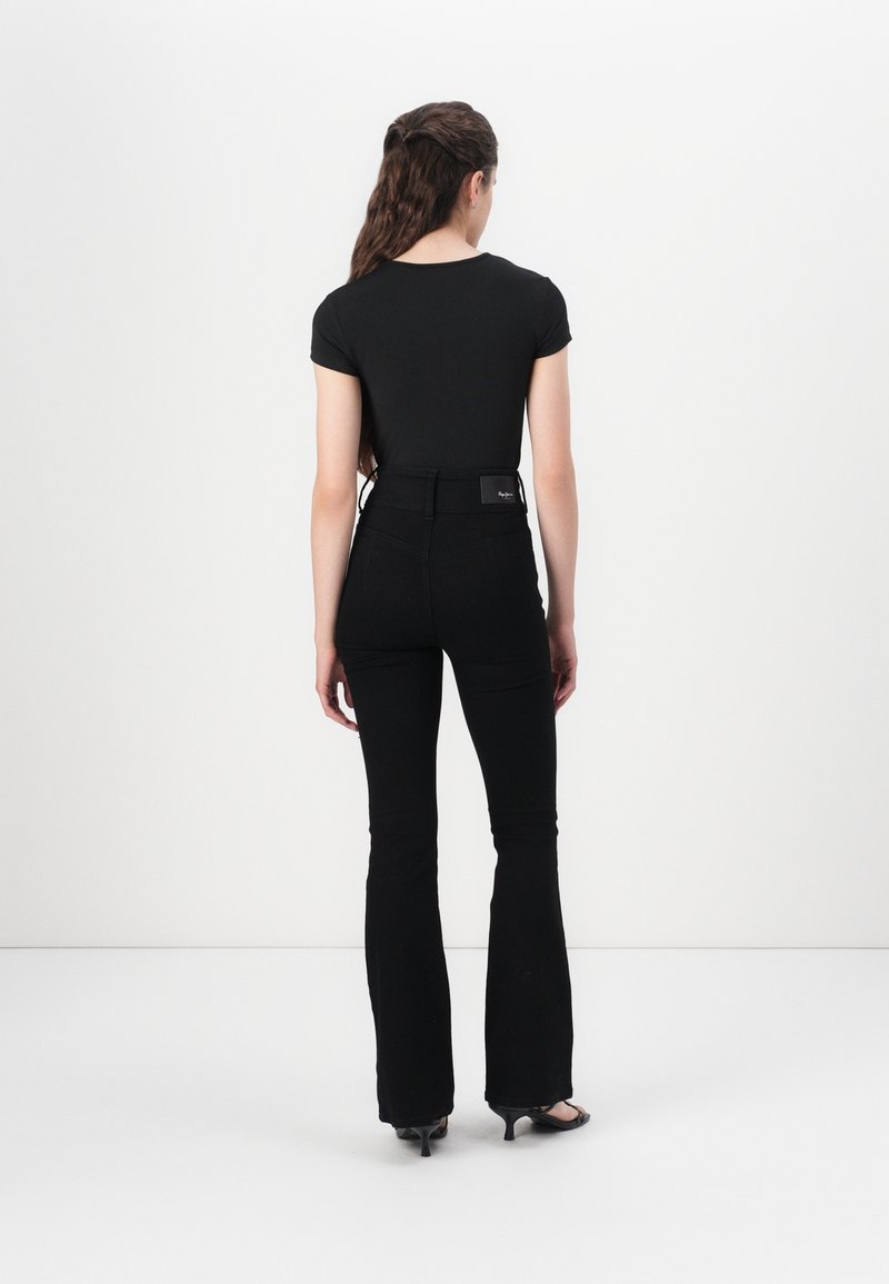 Femme aux longs cheveux ondulés, portant un haut noir cintré à manches courtes, un pantalon noir évasé taille haute et des sandales noires à talons, debout, de dos.