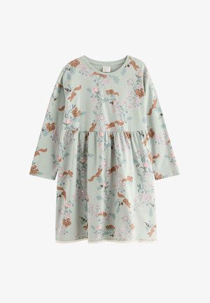 Lille barn med krøllet blondt hår iført en lyseblå kjole med egern- og blomsterprint, hvide sokker og blå sko, der smiler og poserer.