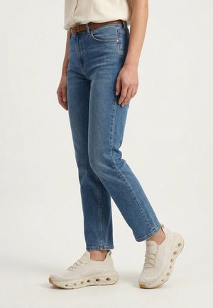 Person iført blå jeans med lige ben, et brunt bælte og beige atletiske sko med strukturerede såler, stående på en lys baggrund.