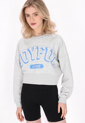 Blonde Frau trägt ein graues Crop-Sweatshirt mit „JOYFUL LEAGUE“ in Blau und schwarze enganliegende Shorts, steht vor weißem Hintergrund.