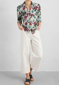 Camicia a bottoni floreale bianca, verde e rossa con maniche lunghe, abbinata a pantaloni a gamba larga color crema e sandali neri.