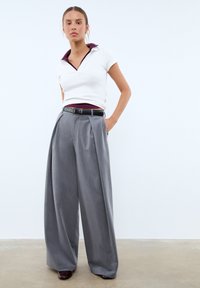 Jeune femme se tenant debout avec une main dans la poche, portant un haut blanc à manches courtes avec un col bordeaux, un pantalon large gris avec une ceinture noire.
