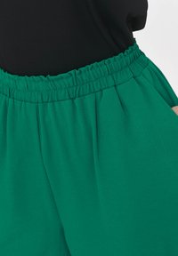 Zielone szorty z tkaniny o elastycznym pasku, charakteryzujące się luźnym krojem i subtelną teksturą. Posiadają kieszenie po bokach i gładkie wykończenie.