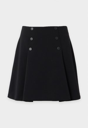 Kobieta w białym, bezrękawnikowym topie z falbankami, czarnej spódnicy w kształcie litery A z guzikami i czarnych balerinkach, stoi z jedną ręką w kieszeni spódnicy.