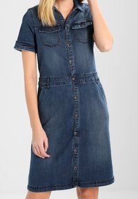 Denim A-linjainen mekko lyhyillä hihoilla, kahdella rintataskulla ja napit edessä, jossa on hieman teksturoitu tumma sininen kangas.