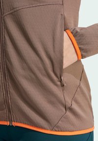 Hnedá vetrovka na zips s oranžovými akcentmi, vyrobená z textúrovanej látky, so bočnými vreckami a priliehavými rukávmi. Dizajn zdôrazňuje pohodlie a praktickosť.