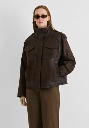 Le modèle porte une veste en cuir marron foncé à col montant et à poches à rabat, un pantalon marron et des lunettes de soleil, sur un fond de studio uni.