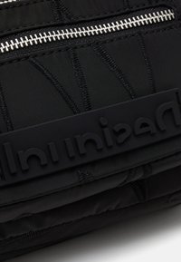 Schwarze Nylon-Tasche mit gestepptem Textil, silbernem Reißverschluss und auffälligem schwarzem Gummilogoelement. Kompaktes Design mit mehreren Fächern.