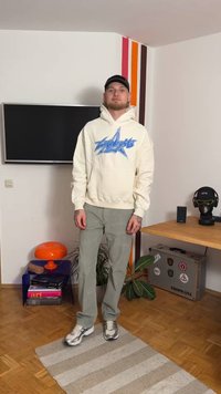 Cremefarbener Hoodie mit blauem Sternlogo, kombiniert mit hellgrünen Hosen. Weiße und silberne Sneaker. Innenraum mit minimaler Einrichtung.
