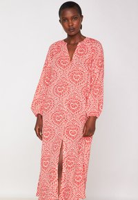 Vrouw in een lange rode en witte gestreepte jurk met een split aan de voorkant, staand voor een effen lichte achtergrond.