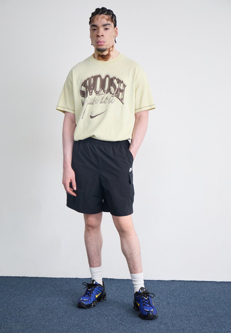 Gelbes Grafik-T-Shirt mit "Swoosh"-Druck, kombiniert mit schwarzen Shorts. Schwarz-blaue Sneaker mit gelben Akzenten. Weiße Crew-Socken.