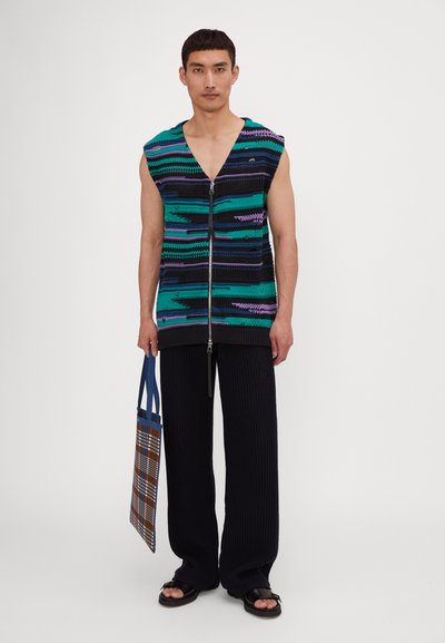 Haut sans manches en tricot avec fermeture éclair, présentant des rayures horizontales vertes, violettes et noires. Associé à un pantalon côtelé noir et un sac fourre-tout à carreaux.