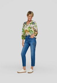 Lächelnde Frau steht mit der Hand in der Tasche, trägt eine mehrfarbige gemusterte Bluse, blaue Jeans mit Stern-Patches und weiße Sneakers.