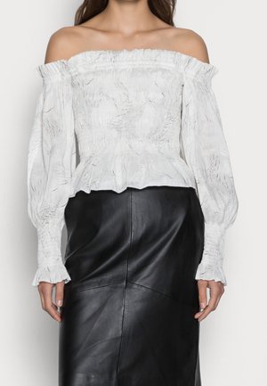 Femme portant un chemisier blanc smocké à épaules dénudées avec manches longues et une jupe en cuir noire taille haute, debout devant un fond uni.