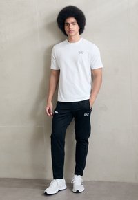 Fehér, rövid ujjú póló EA7 logóval, fekete, szűkített szabású tréningnadrággal, amelyen EA7 márkaelnevezés található. A modellen fehér sportcipők vannak.