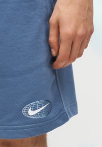 Χέρι που ακουμπάει δίπλα σε μπλε σορτσάκια με λευκό λογότυπο της Nike πάνω σε σχέδιο υδρογείου κοντά στο τελείωμα.