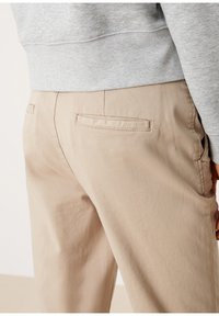 Detailní záběr na zadní kapsu béžových kalhot a část šedého svetru, ukazující švy a texturu látky.