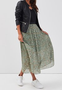 Jupe maxi plissée avec un motif floral doré, base verte, tissu transparent ; associée à une veste en cuir noire et des baskets blanches.