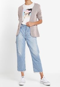 Stickad cardigan i ljusbrunt, öppen design fram; grafisk vit t-shirt; avslappnade ljusblå jeans med fransar i nederkant; vita sneakers.