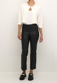 Weiße cropped Strickjacke mit 3/4-Ärmeln, kombiniert mit glänzenden schwarzen Faux-Lederhosen. Schwarze Slipper mit goldenen Akzenten runden den Look ab.
