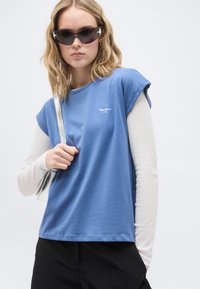 Νεαρή γυναίκα φορώντας μπλε αμάνικο τοπ πάνω από λευκό μακρυμάνικο, μαύρο παντελόνι, γυαλιά ηλίου με διακόσμηση αστέρια και κρατώντας ασημένια τσάντα ώμου.