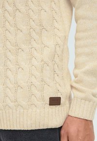 Pulover crem din tricot cu cablu, care prezintă modele texturate, manșete cu nervuri și un mic eticheta din piele maro. Țesătură moale cu un design voluminos.