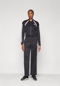 Svart zip-up träningsoverall med rosa detaljer, som har vita ränder längs ärmarna och byxorna. Bärs med svarta träningsskor.