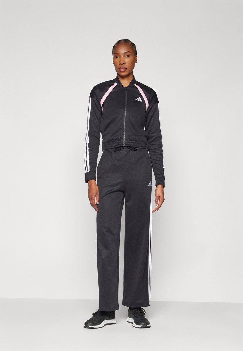 Svart zip-up träningsoverall med rosa detaljer, som har vita ränder längs ärmarna och byxorna. Bärs med svarta träningsskor.