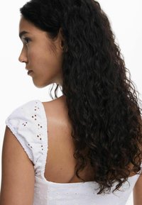 Valge kleit, millel on broderiidiga lillemuster, lühikesed pahklaiustavad varrukad ja ümar tagumine osa. Kleit on valmistatud tekstuuritud kangast ning selle kandjal on lainelised tumedad juuksed.