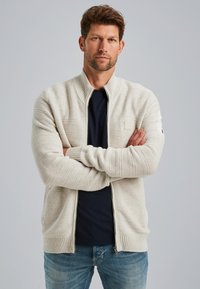 Lichtbeige ritscardigan van textuurgebreid materiaal, met ribgebreide boorden en onderkant, en een subtiele logopatch op de borst. Gemaakt in combinatie met een donker shirt eronder.