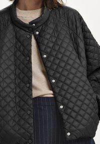 Person iført en sort kviltet jakke med trykknapper over en beige sweater og marineblå stribede bukser mod en ensfarvet baggrund.