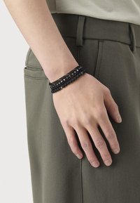 Mand hånd hviler på lommen på olivengrønne bukser, iført tre sorte perle- og snorearmbånd på håndleddet.