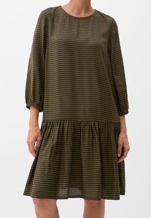 Kobieta ubrana w oliwkowo-zieloną i czarną kraciastą sukienkę z długim rękawem, o obniżonej talii i marszczonym dole, stojąca na białym tle.