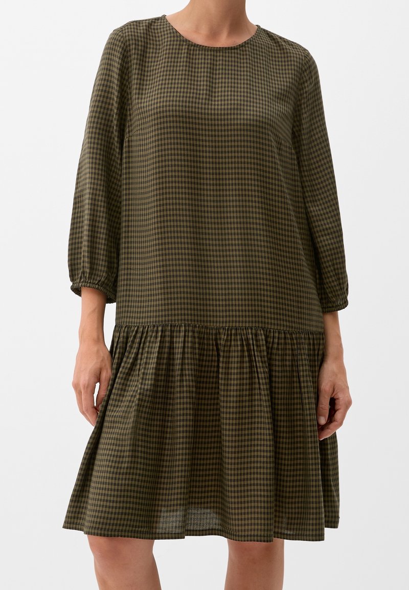 Femme portant une robe à manches longues à carreaux vert olive et noir, avec une taille basse et une jupe froncée, debout devant un fond blanc.